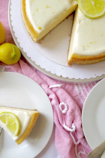 A velvety smooth and luscious looking lemon cheesecake on a pink table cloth. A slice of cheese cake is next to it on a side plate and there are fresh lemons beside it.