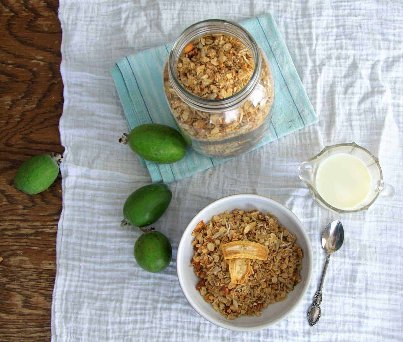 A breakfast bowl of feijoa granola, alongside a jug of milk, a spoon, fresh feijoas, and a jar of feijoa granola.