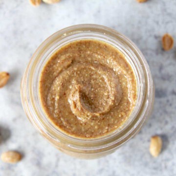 A top view of homemade peanut butter in a glass jar. There is a beautiful creamy swirl in the peanut butter and roasted peanuts scattered around the jar.