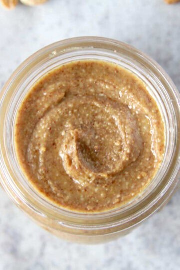 A top view of homemade peanut butter in a glass jar. There is a beautiful creamy swirl in the peanut butter and roasted peanuts scattered around the jar.