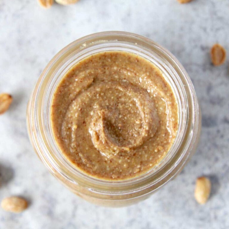 A top view of homemade peanut butter in a glass jar. There is a beautiful creamy swirl in the peanut butter and roasted peanuts scattered around the jar.