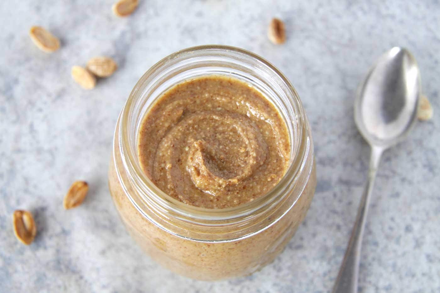A marble background with a glass jar filled with homemade peanut butter swirled inside the jar. A spoon and roasted peanuts are scattered around the jar.