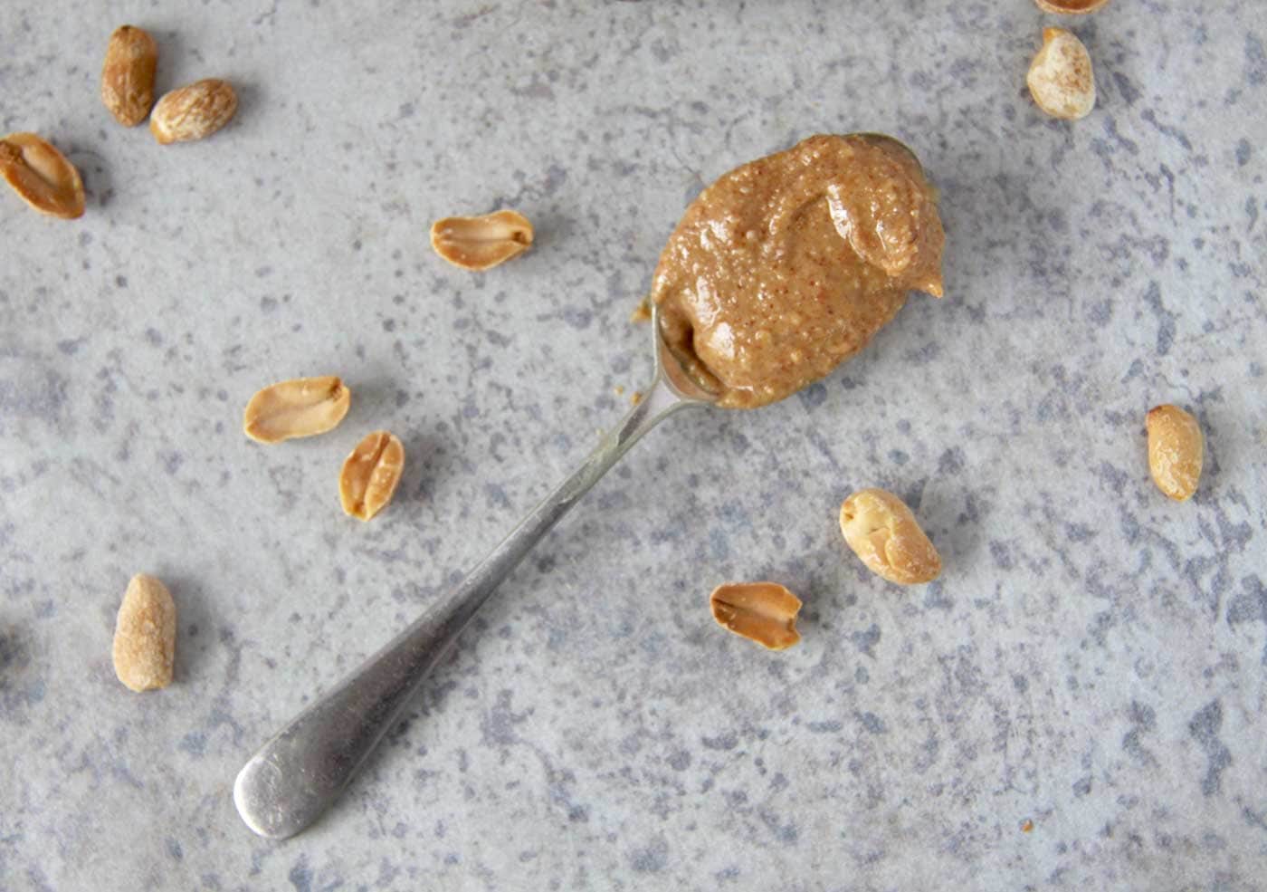 A spoon of freshly homemade peanut butter sitting on a marble bench with roasted peanuts scattered around it.