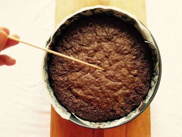 A chocolate cake in a cake tin lined with baking paper. A hand is holding a bamboo skewer in the middle of the cake to see if it is done.