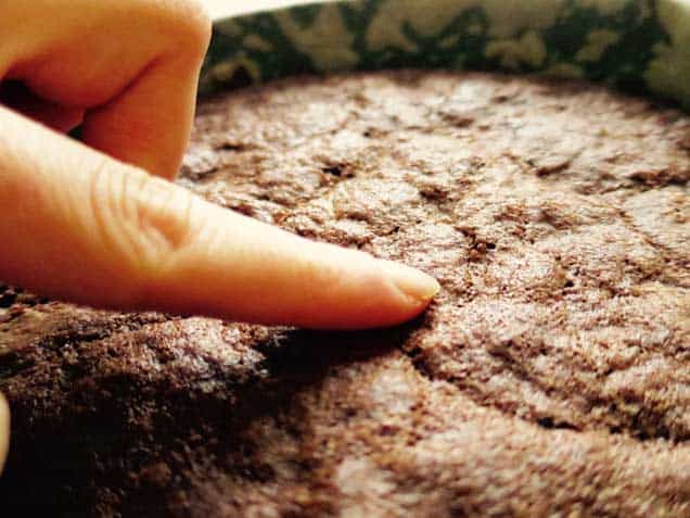 A finger pressing gently on a chocolate cake still in it's tin. Checking to see if it is done baking.