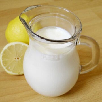 A small glass jug of buttermilk with lemon halves on the side on a table.