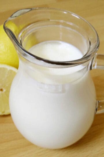A small glass jug of buttermilk with lemon halves on the side on a table.