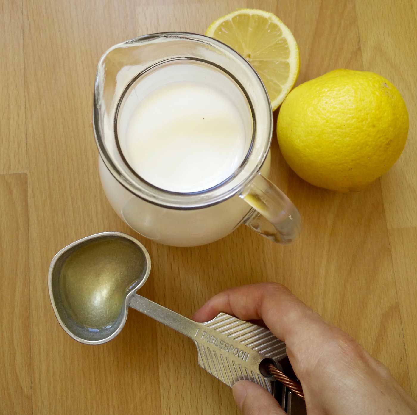 A jug of milk with a hand holding a heart shaped measuring spoon filled with lemon juice, ready to pour into the milk.