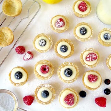 Mini lemon curd tarts on a bench with a glimpse of a few empty shortbread cases that still need filling. Fresh raspberries and blueberries are used to decorate the tarts.