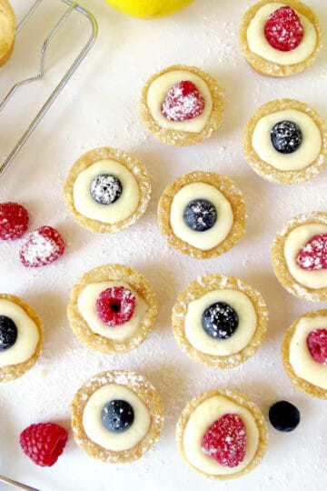 Mini lemon curd tarts on a bench with a glimpse of a few empty shortbread cases that still need filling. Fresh raspberries and blueberries are used to decorate the tarts.