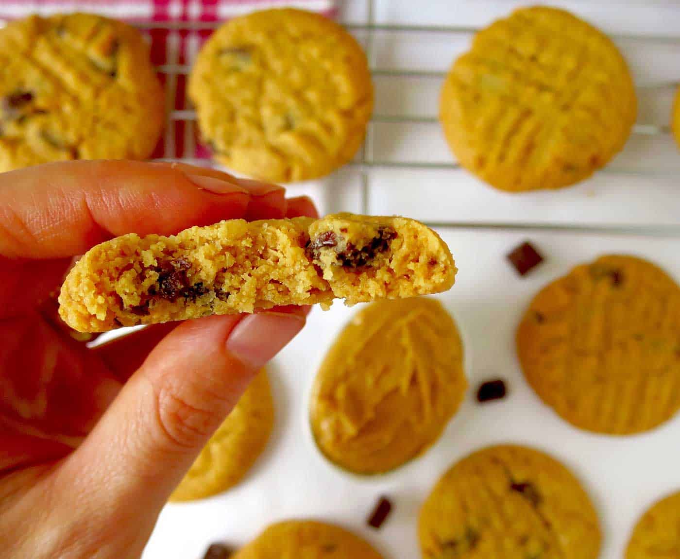 A hand holding a peanut butter cookie which has been bitten into. In the background there are more cookies on a white table and some cooling on a cooling rack.