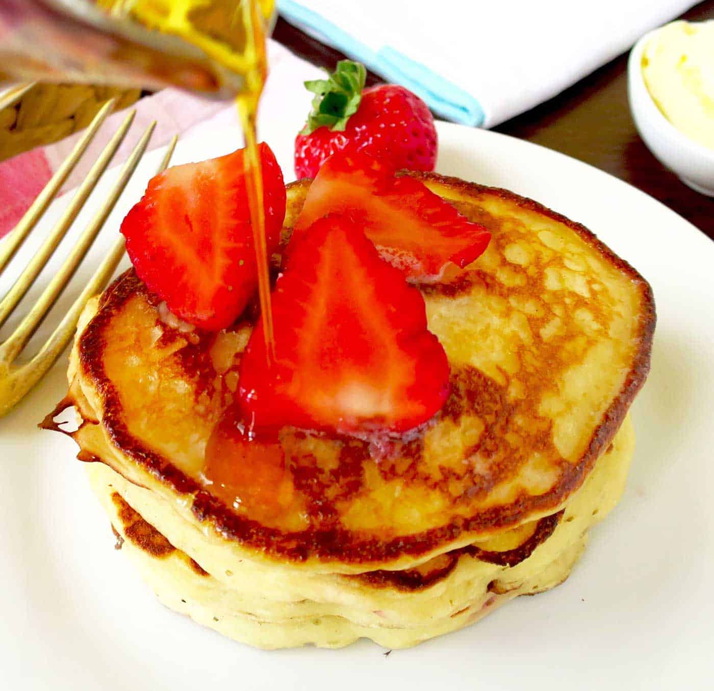 A generous helping of pancakes stacked on a side plate with fresh strawberries on top and a hand pour maple syrup on top.