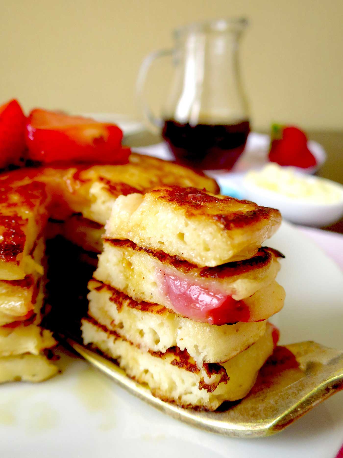 A close up shot of a fork with four layers of pancakes cut into a small wedge to eat. Maple syrup can be seen in the background.