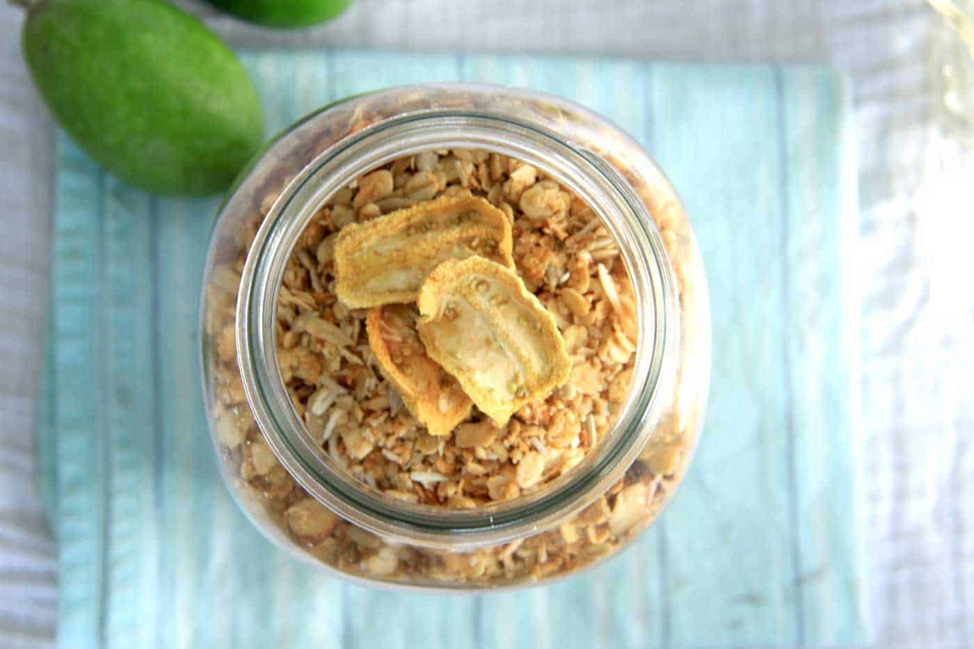 A top view looking into a large jar filled with feijoa granola. Three slices of dried feijoas are on top.