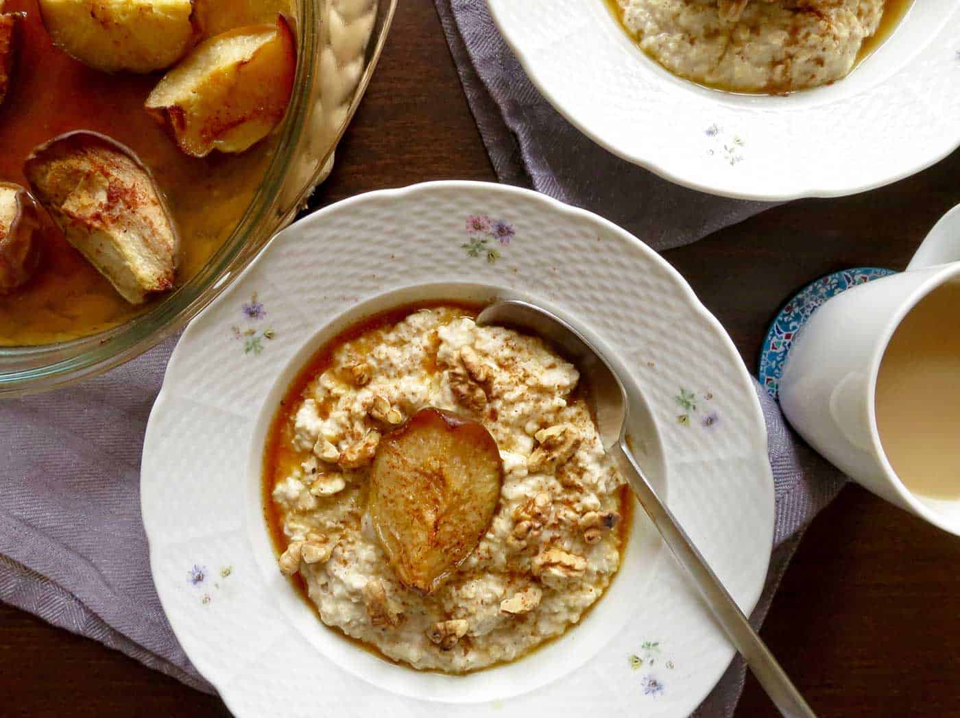 A vintage bowl sitting on a linen blue tea towel filled with creamy vanilla porridge topped with a maple roasted pear quarter and walnuts sprinkled on top. A spoon is at the ready for scooping the first mouthful. Another bowl of porridge can just be seen poking in at the edge of the image. A cup of tea is also on the table and a glimpse of the dish with roasted pears can just be seen also.