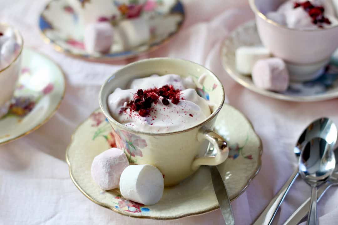 Pink berry ambrosia with berry powder sprinkled on top served in a delicate vintage tea cups. Two marshmallows on the saucer.