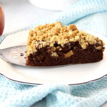 A piece of moist chocolate apple crumble cake with crumble topping. There are pieces of juicy apples studded through the cake. It is resting on a cake server and being plated onto a side plate. There is a blue tea towel underneath the plate.