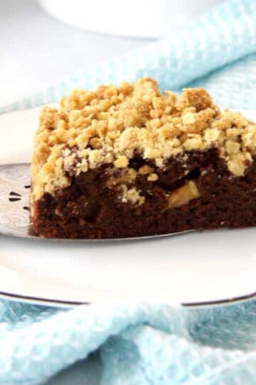 A piece of moist chocolate apple crumble cake with crumble topping. There are pieces of juicy apples studded through the cake. It is resting on a cake server and being plated onto a side plate. There is a blue tea towel underneath the plate.