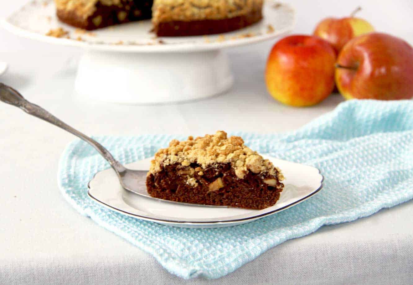 A chocolate apple crumble crumble cake slice on a side plate with cake slice still underneath it. The crumble topping is crunchy in texture and thick. There are pieces of apple studded in the moist looking chocolate cake.