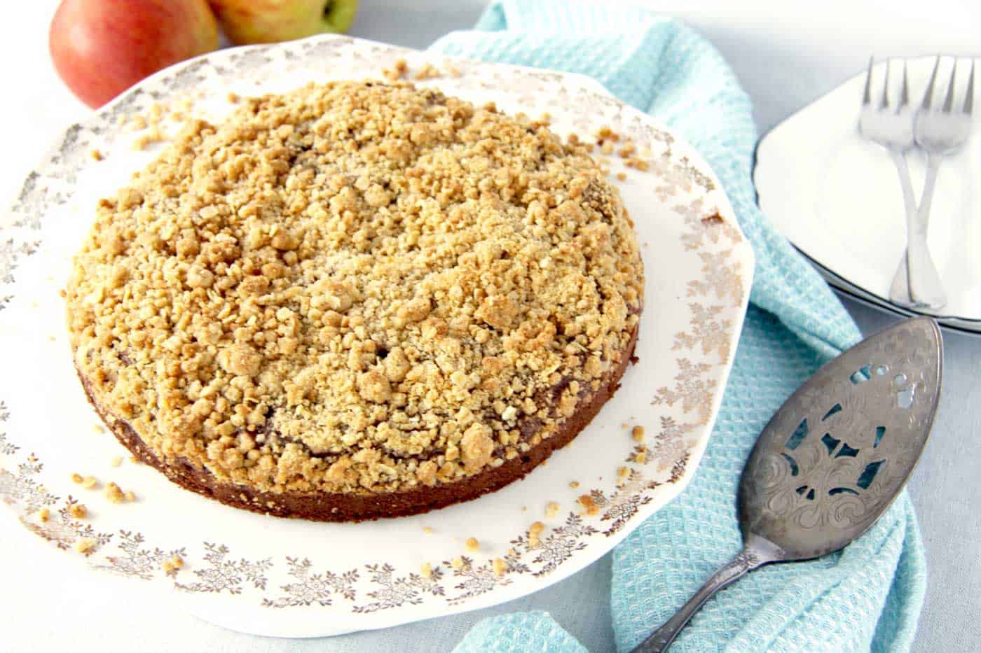 A chocolate apple crumble situated on a cake stand with side plates, forks, fresh apples and a cake server on the side. A blue tea towel is underneath which makes the cake really pop.