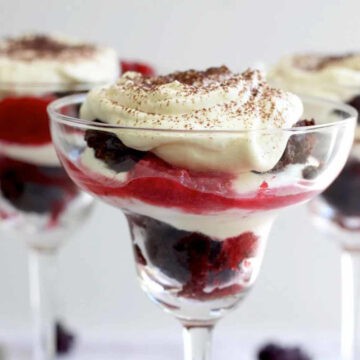 Three margarita glasses two at the back and one up close. All filled with berry chocolate trifles and topped with a lashing of whipped cream and sprinkled with sifted cocoa powder. The layers of chocolate cake, berry compote and cream really stand out.