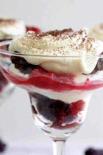 Three margarita glasses two at the back and one up close. All filled with berry chocolate trifles and topped with a lashing of whipped cream and sprinkled with sifted cocoa powder. The layers of chocolate cake, berry compote and cream really stand out.