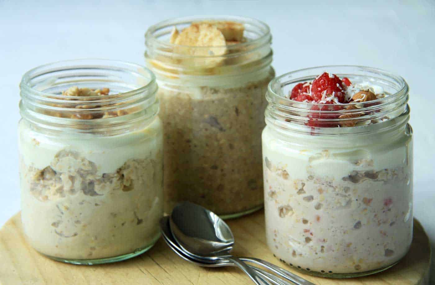 Three jars of overnight oats sitting o na board with spoons. One is berry, one peanut butter and the other banana bread flavoured.