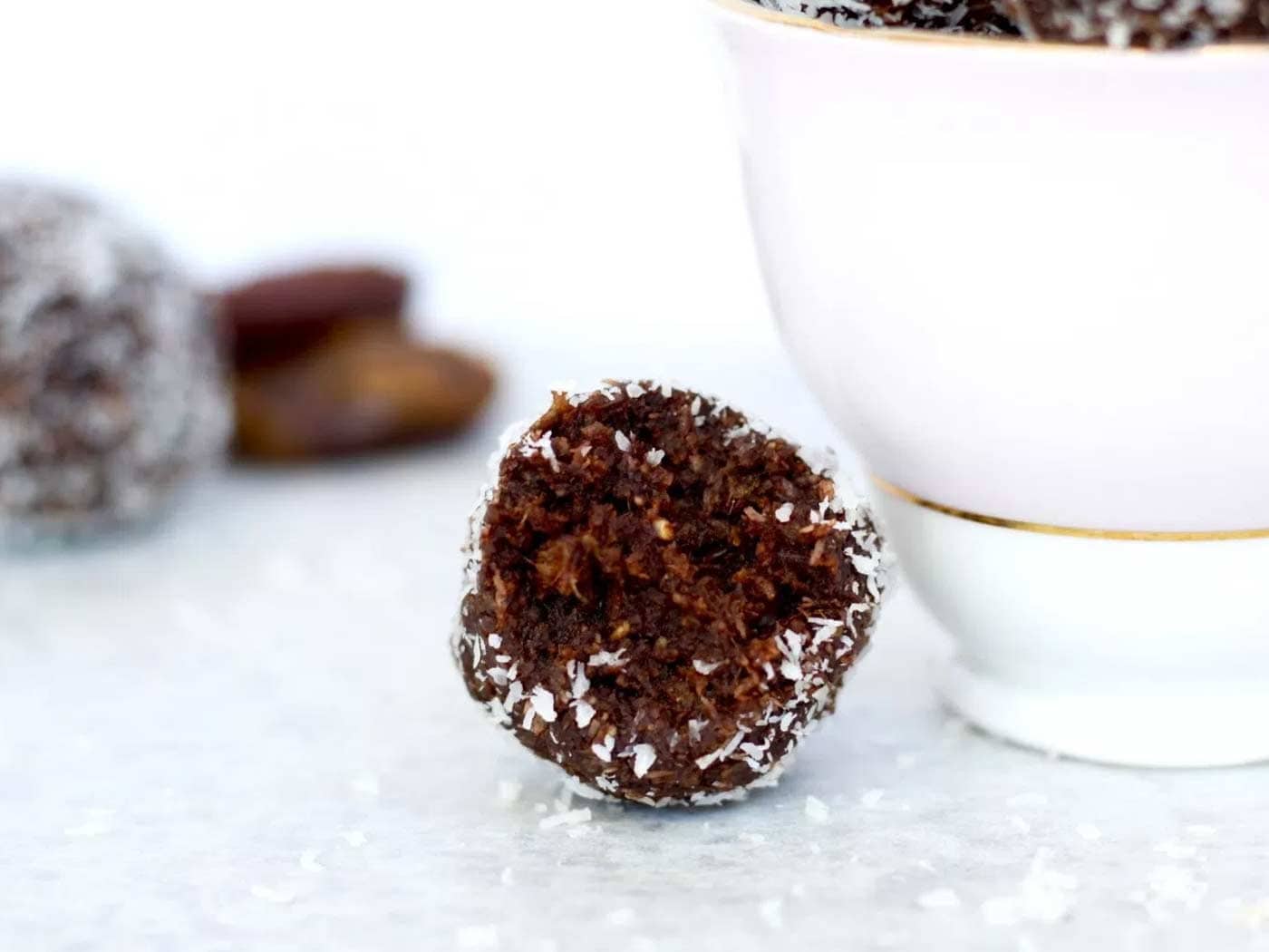 Close up of bliss ball which has been cut in half sitting next to a pink tea cup which has more vegan bliss balls in it.