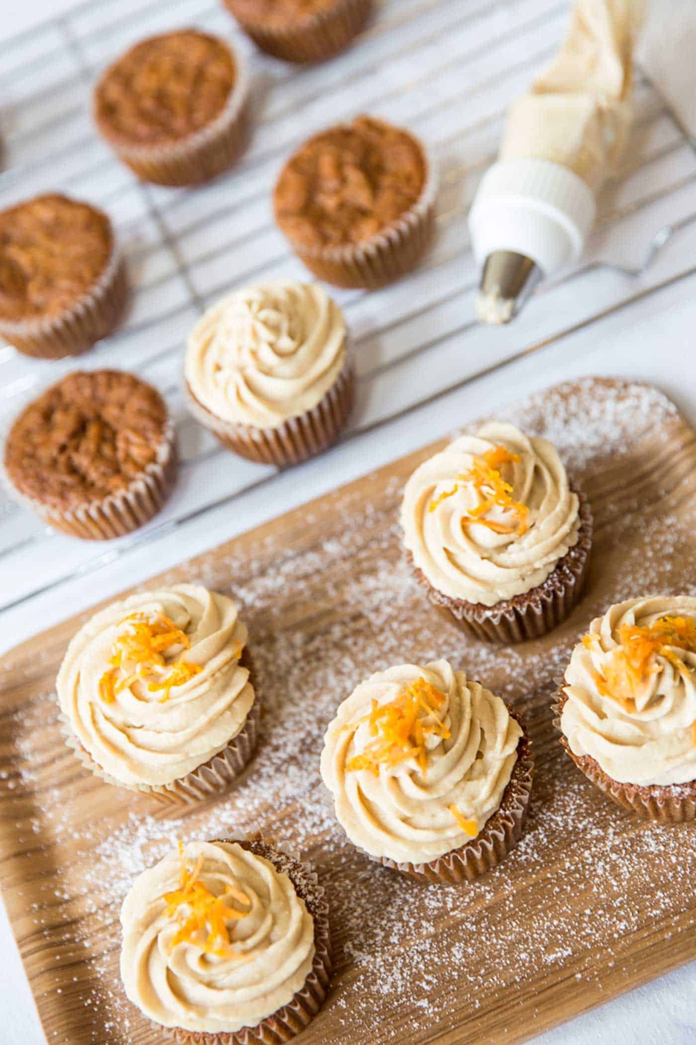 Delicious brown sugar cream cheese frosting carrot cupcakes on a tray with a few un-iced cupcakes on a cooling rack nest to them. The iced cupcakes are decorated with orange zest.