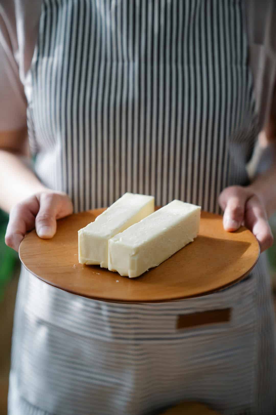 A person with a striped apron holding a dish that has two sticks of butter on it.