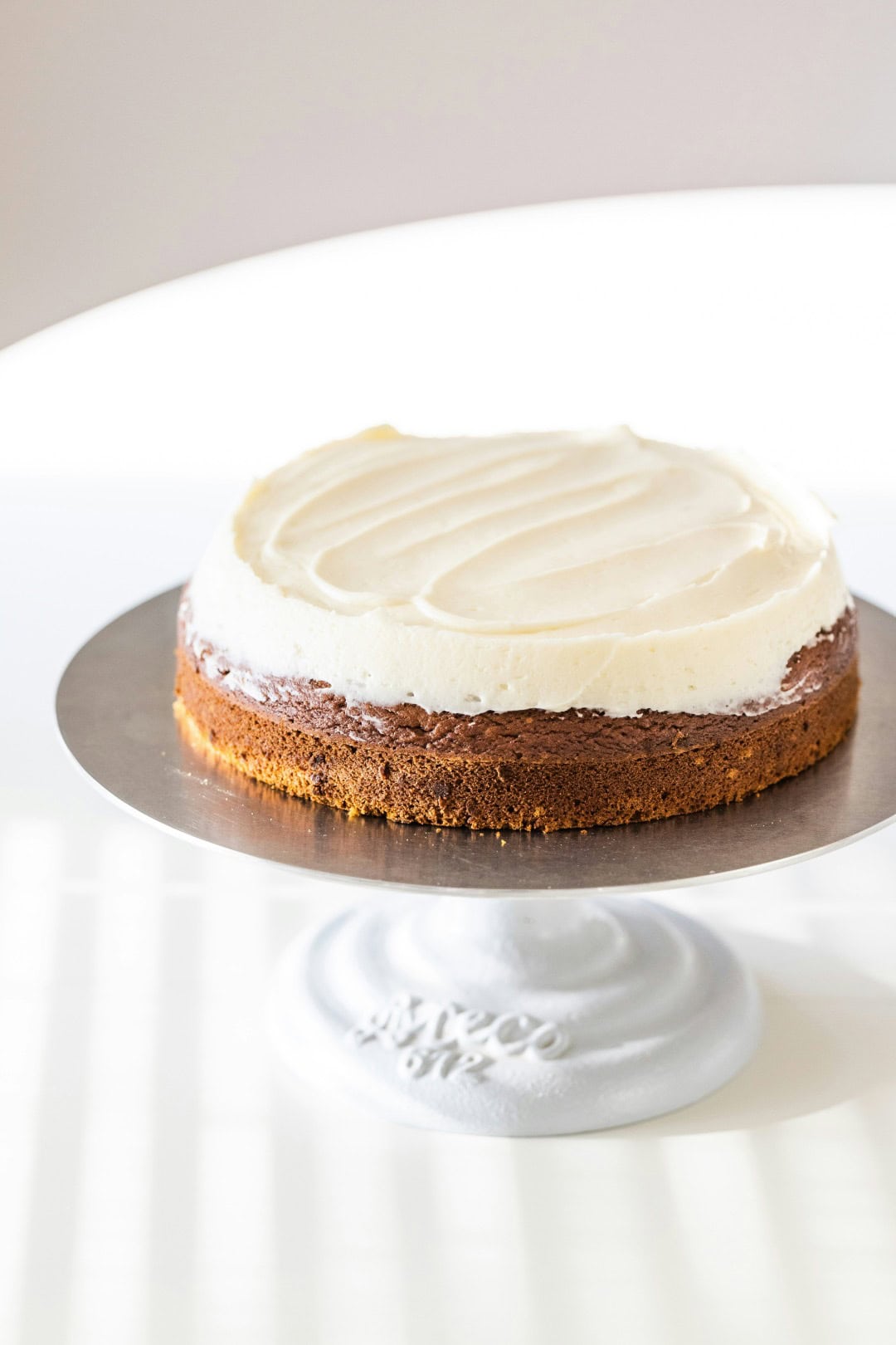 Perfect and luscious white cream cheese frosting iced on a carrot cake. The cake is sitting on a silver cake stand which is one a white table.