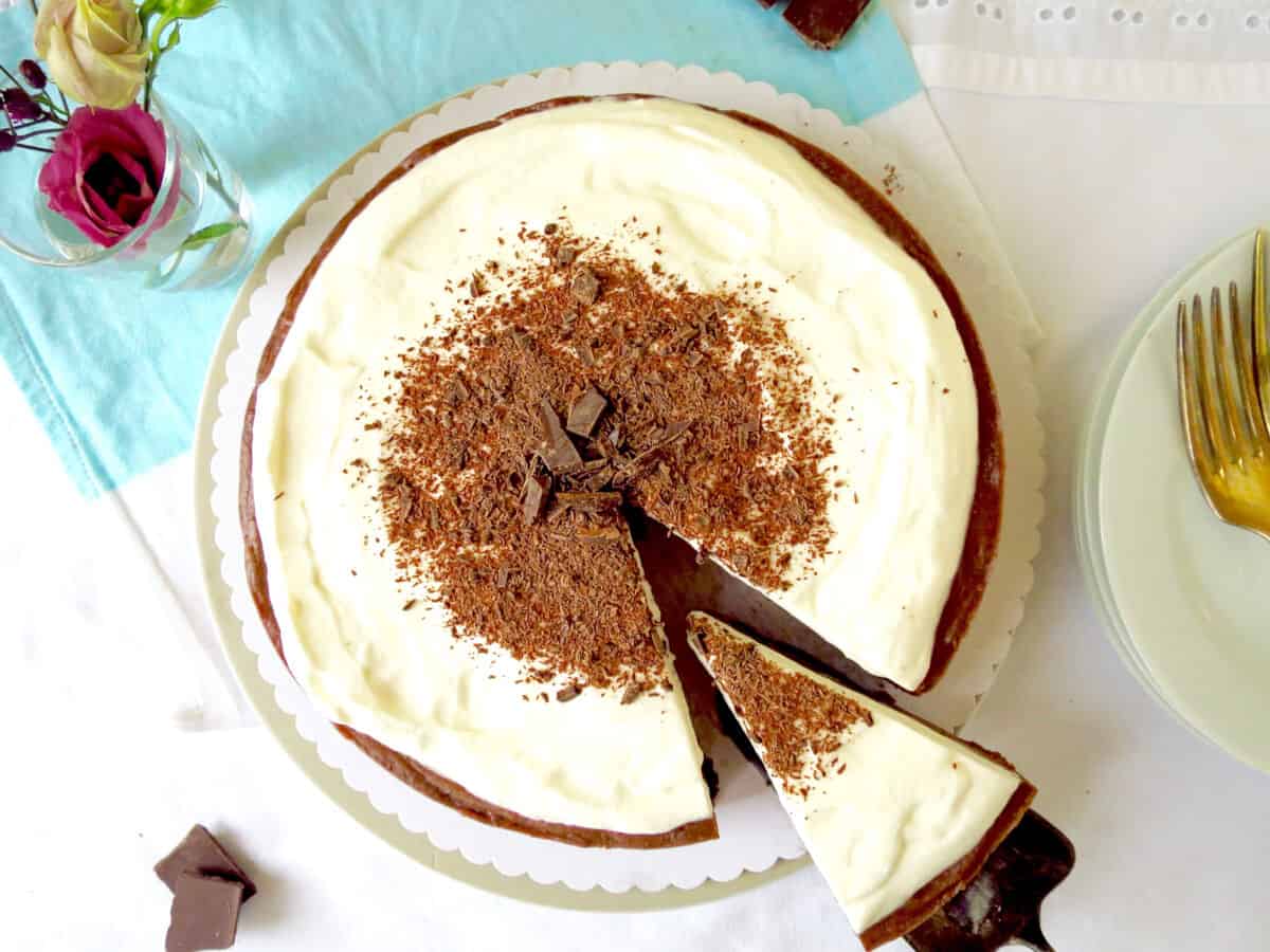 A top view of the velvet chocolate cheesecake. It has a creamy sour cream topping which is decorated with chocolate shards. A slice has been cut and about to be served on a plate nearby.