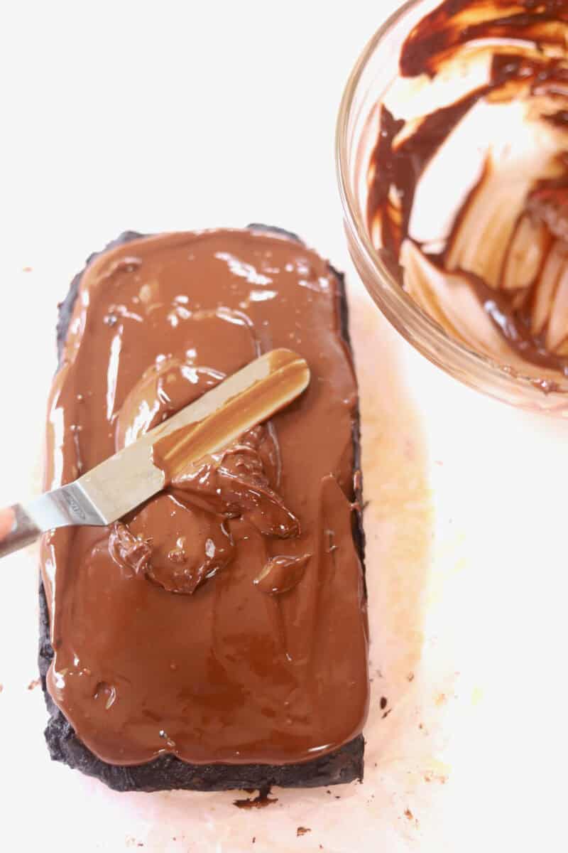 A small off-set spatula spreading chocolate ganache on top of the chocolate vegan brownie before cutting into pieces. An empty bowl of ganache is next to it.