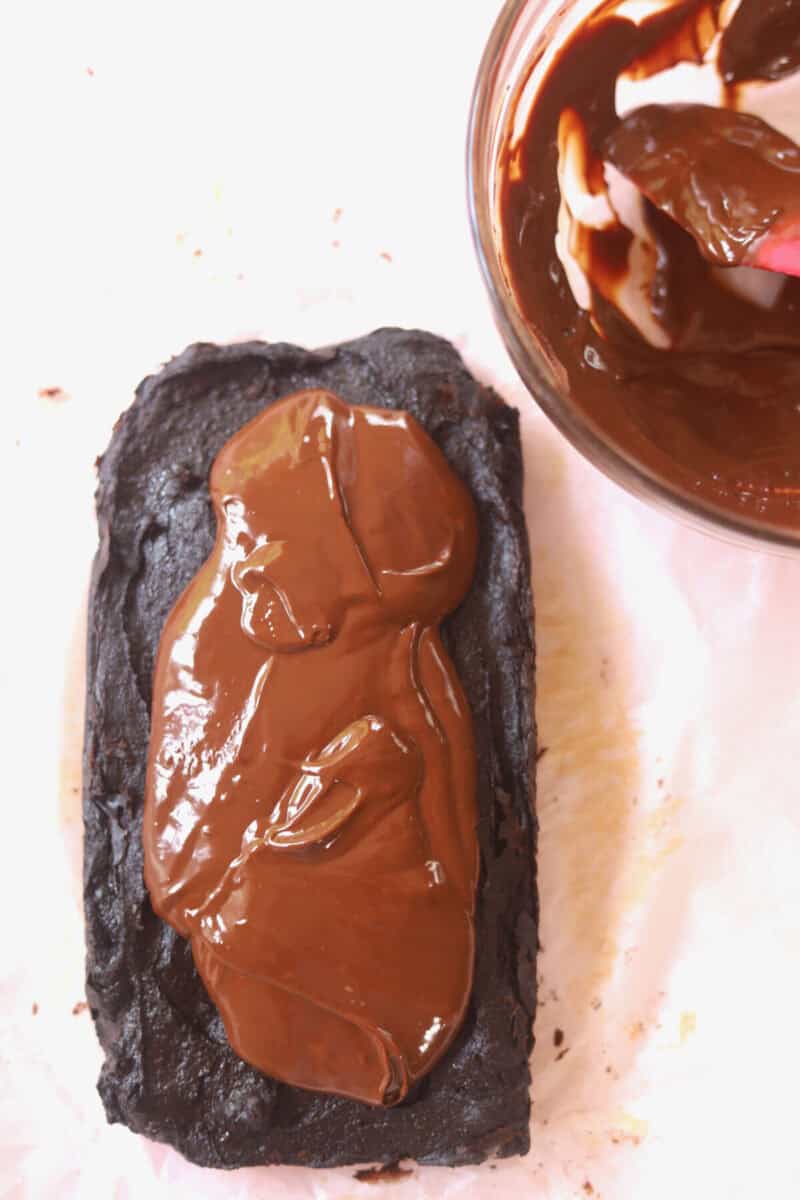 Chocolate ganache slathered on top of the vegan chocolate brownie. A spatula and remaining chocolate ganache is in a bowl next to it.