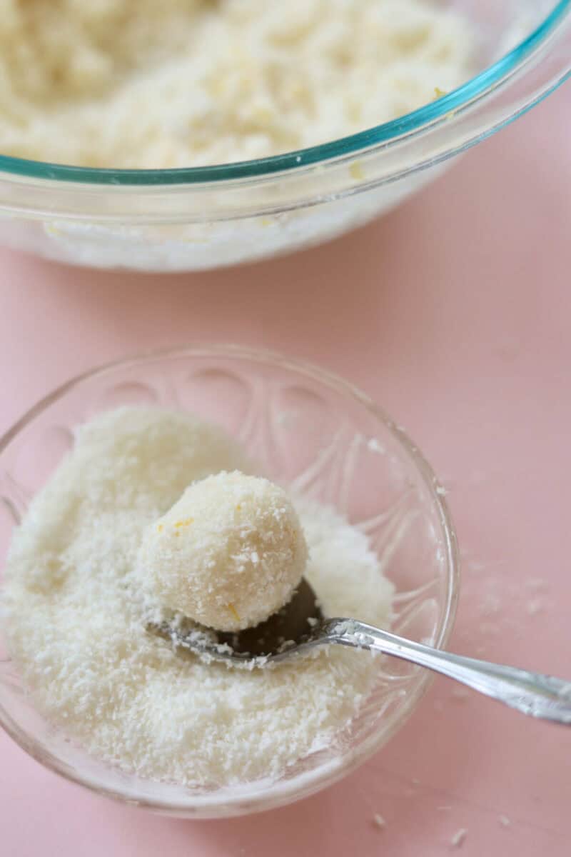 A lemon coconut truffle which has just been rolled in coconut with a spoon in a bowl of coconut.