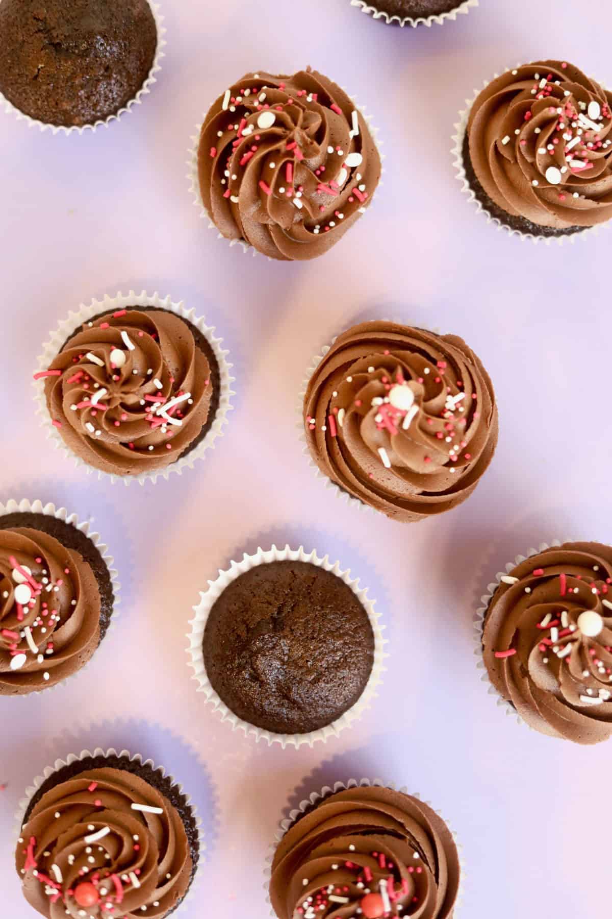 An array of cupcakes with a purple background. Some are decorated with frosting and sprinkles, and some are plain.