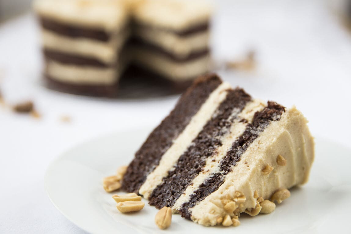 A slice of moist chocolate layer cake with peanut butter buttercream. The rest of the cake is blurred in the background.