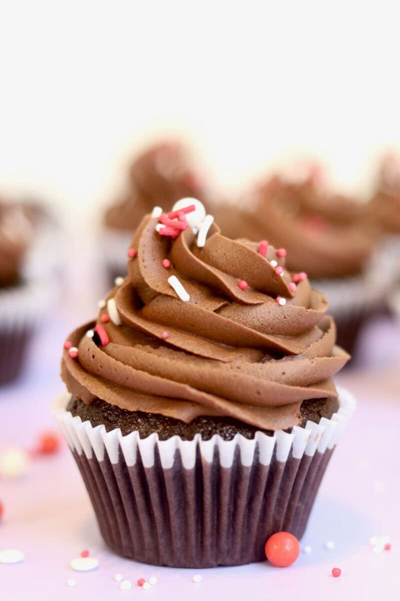 A divine and beautiful looking decadent chocolate cupcake in the foreground. It has been frosted with a perfect buttercream swirl and topped with red and white sprinkles. There are more cupcakes in the background which have been blurred.