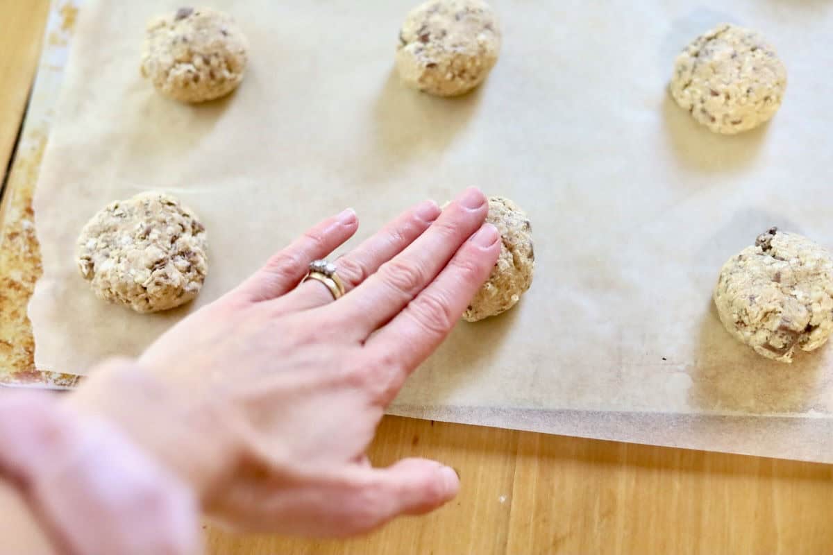 Lactation cookie dough rolled into balls on a baking tray lined with baking paper. A hand is gently pressing on the balls to flatten them slightly.