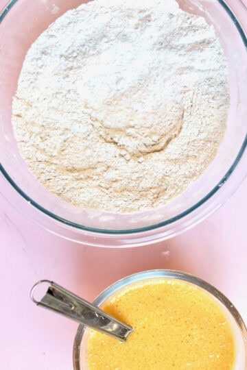 Two glass bowls on a pink board. One larger bowl has dry ingredients for a muffin recipe and the other has wet ingredients. In preparation for baking using the muffin method.