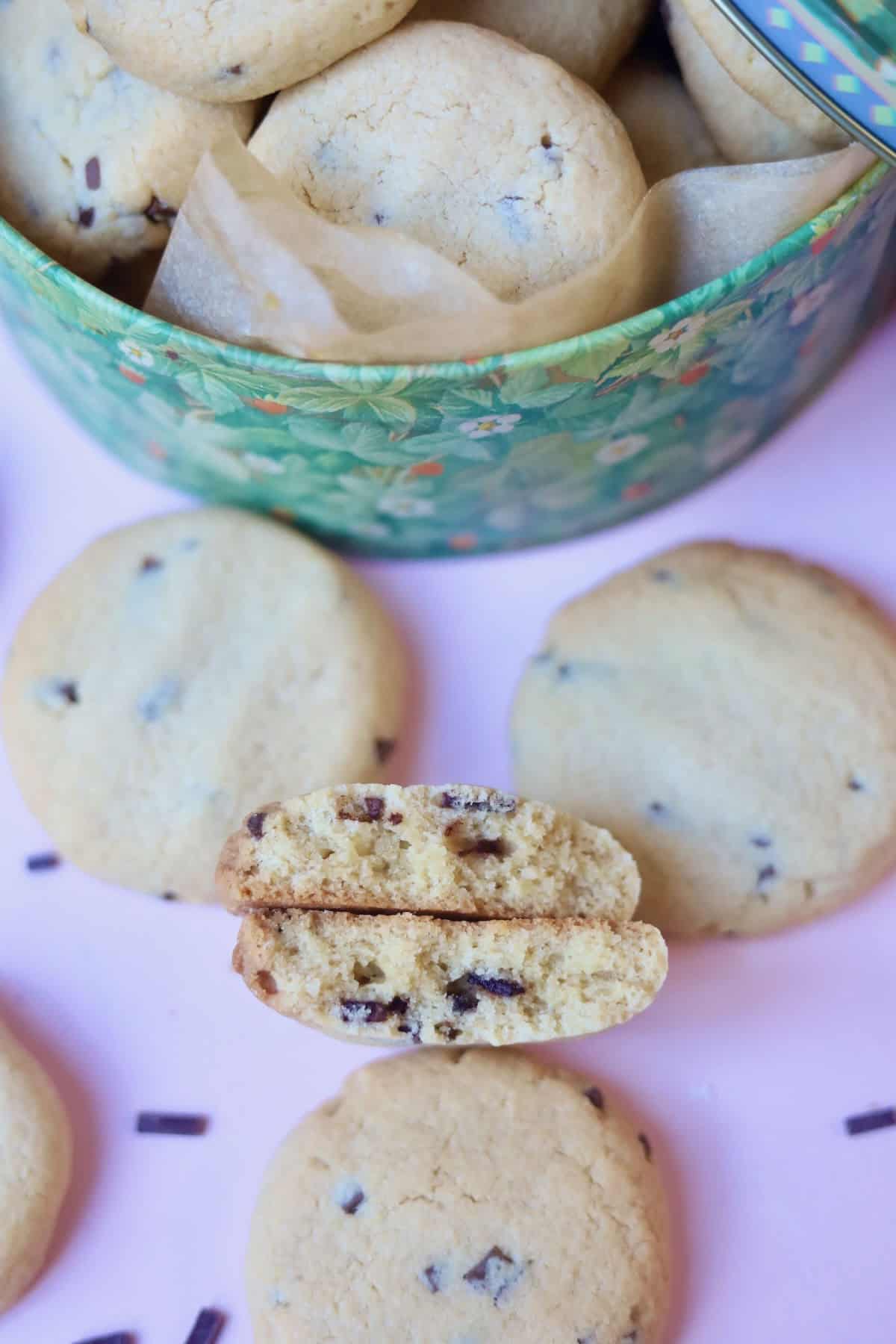 A cookie tin filled with cookies and some next to it, show casing a cookie which has been broken in half to reveal the tender texture.