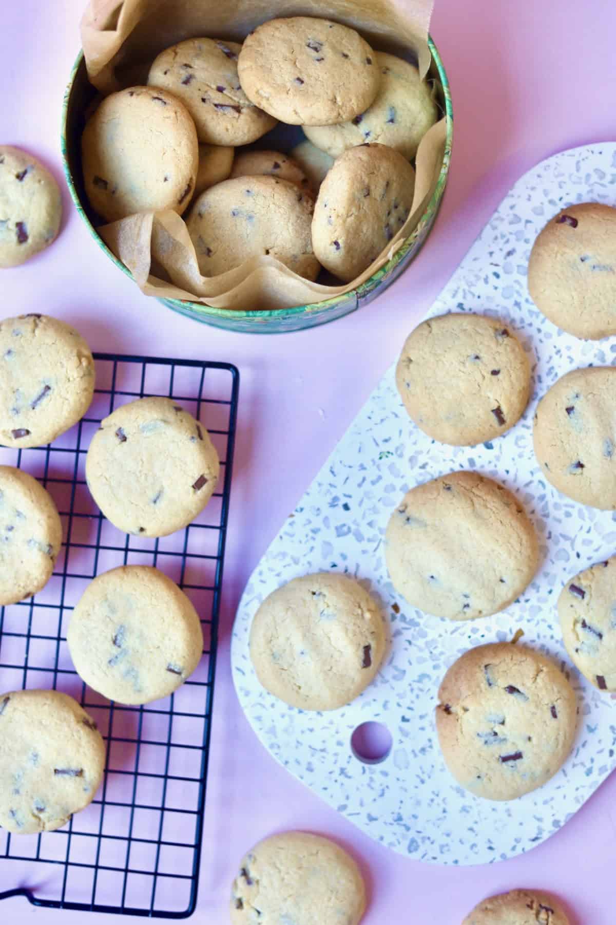 A cookie tin filled with cookies, a cooling rack filled with cookies and a white board, displayed with more cookies.