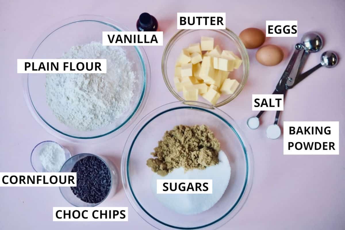 All ingredients for chocolate chip cookie recipe flat laid on a pink background with each corresponding word for each ingredient.