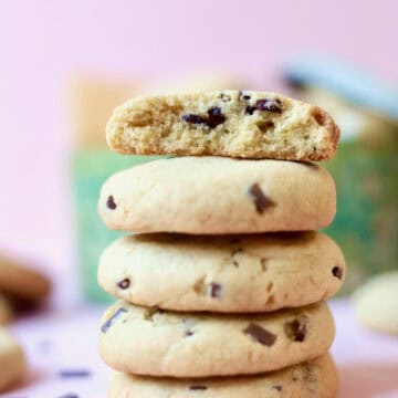 A stack of chocolate chip cookies with a cookie tin in the background filled with more cookies.
