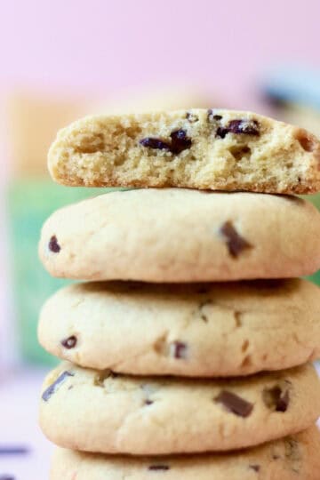 A stack of chocolate chip cookies with a cookie tin in the background filled with more cookies.