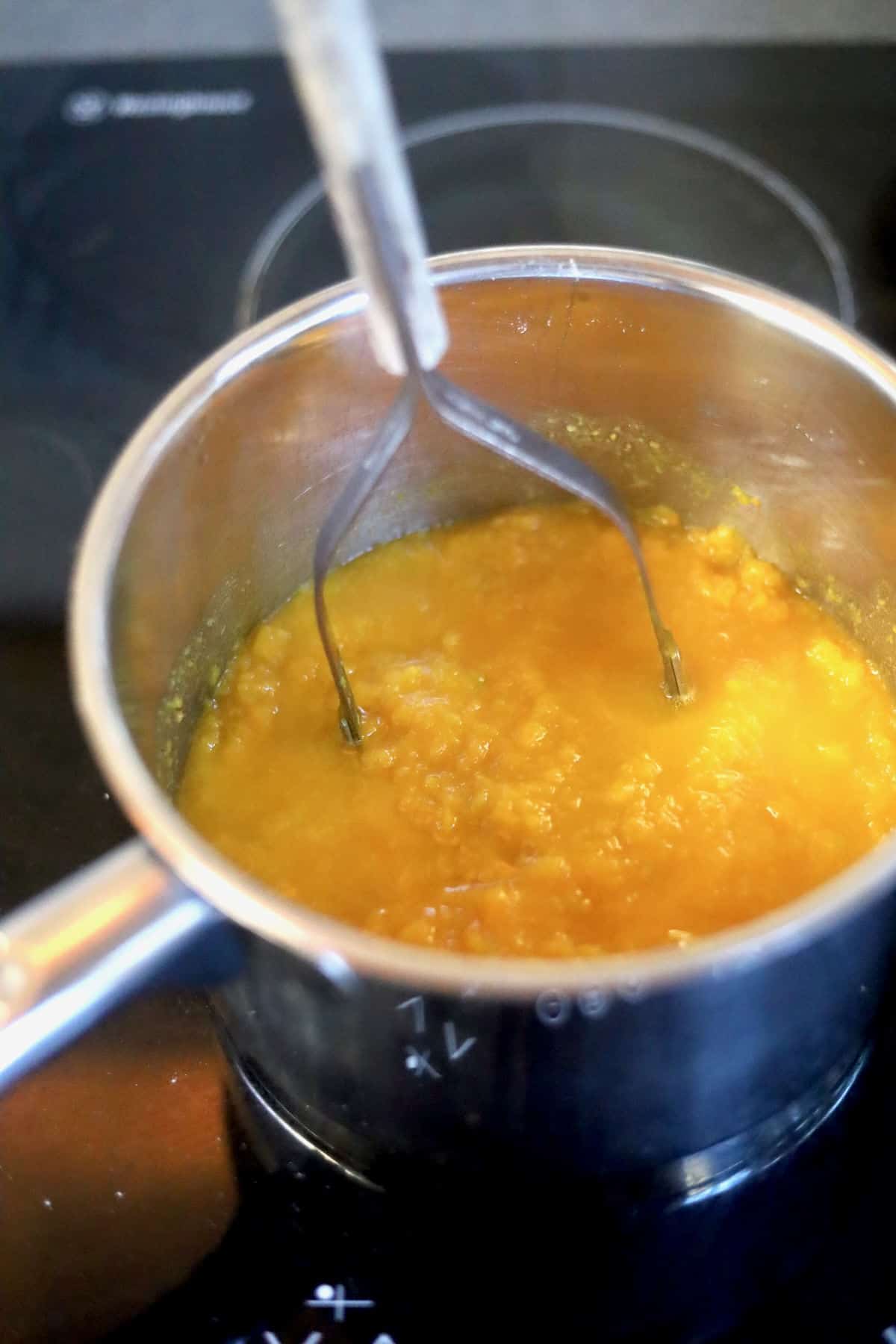Cooked pumpkin in a saucepan with a potato masher mashing it.