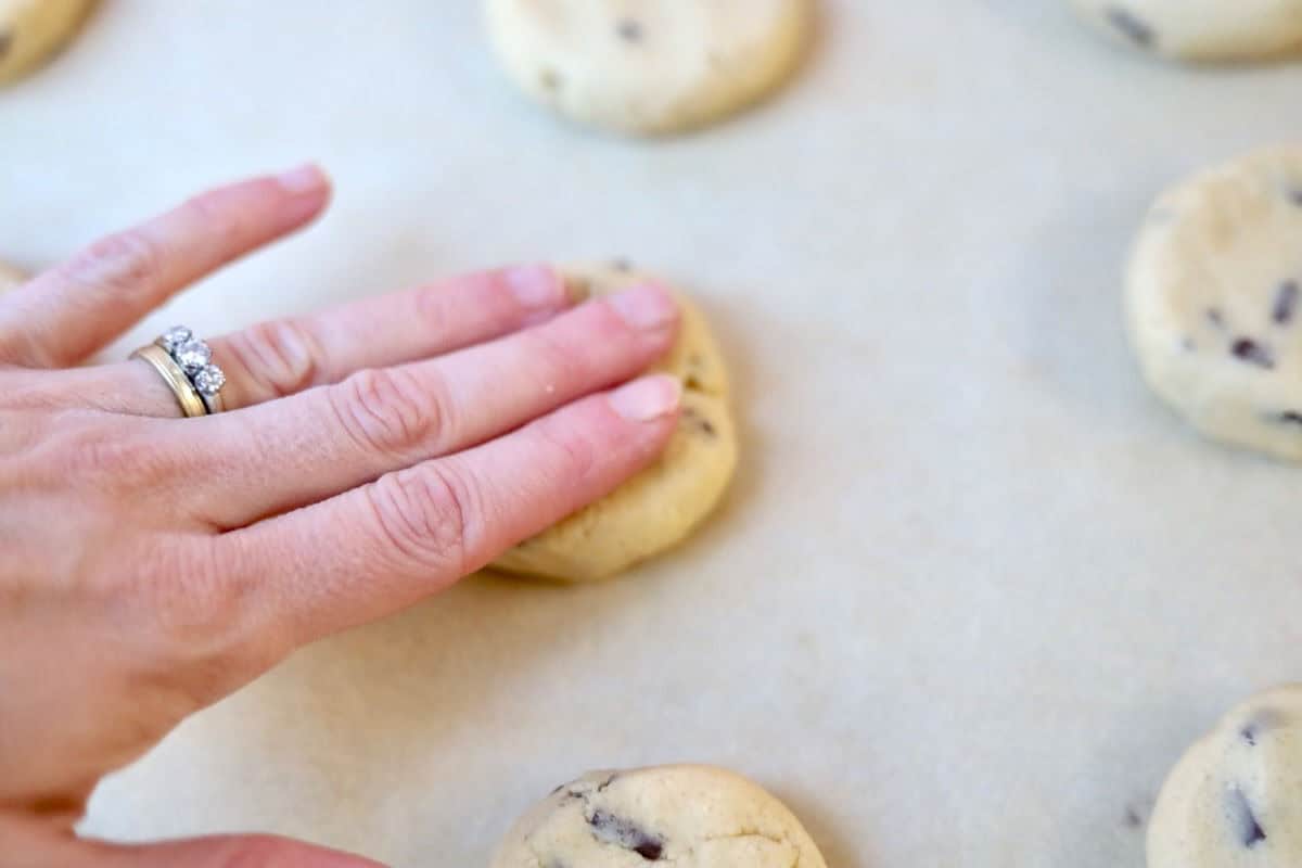 A hand is gently pressing on the cookie dough ball to flatten it slightly.